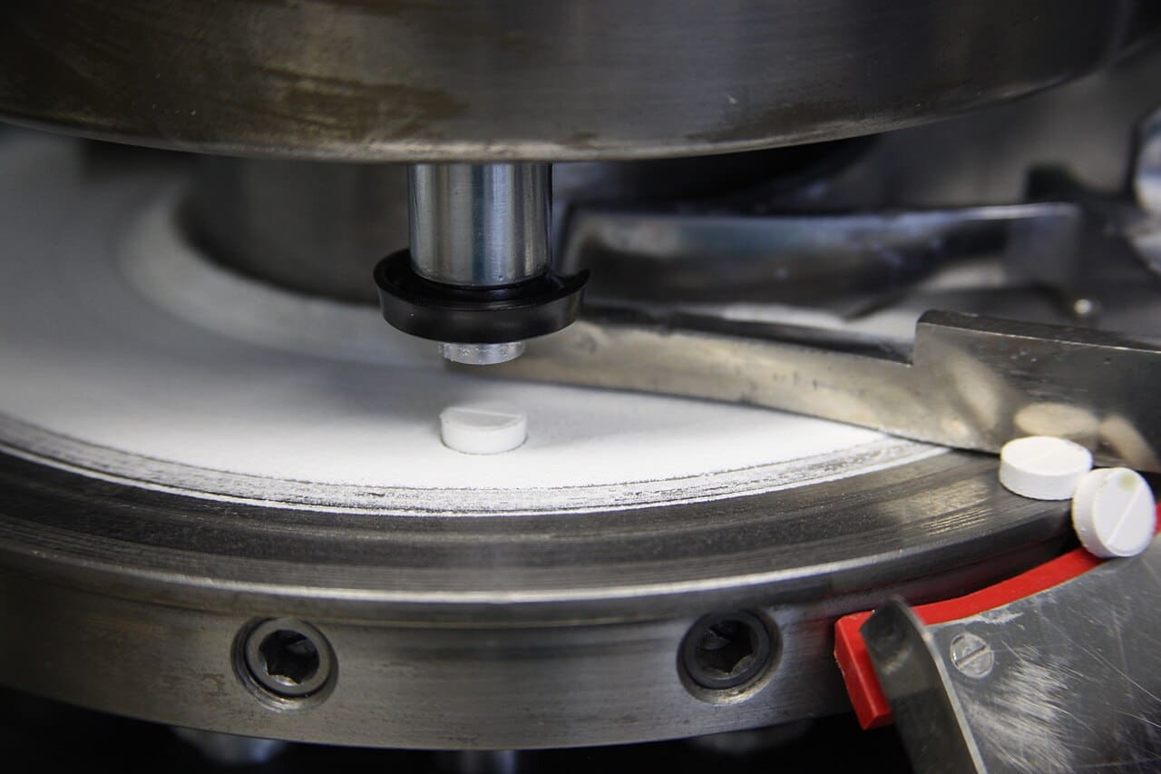 Close-up of a rotary tablet press: an upper punch above the die table with finished tablets